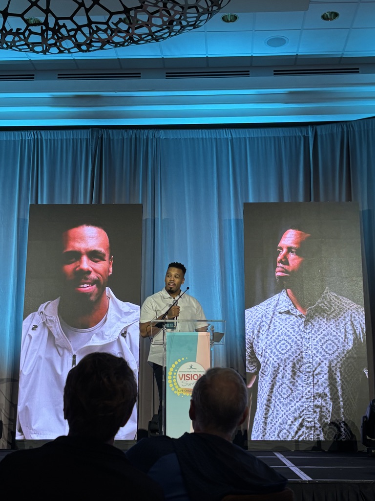 Rolando “Roe” Ruiz speaking from the podium at a summit, his portraits projected on screens behind him